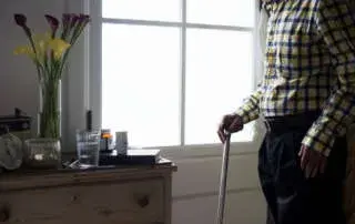 Pessoa idosa usando bengala dentro de casa, com objetos de uso diário próximos.