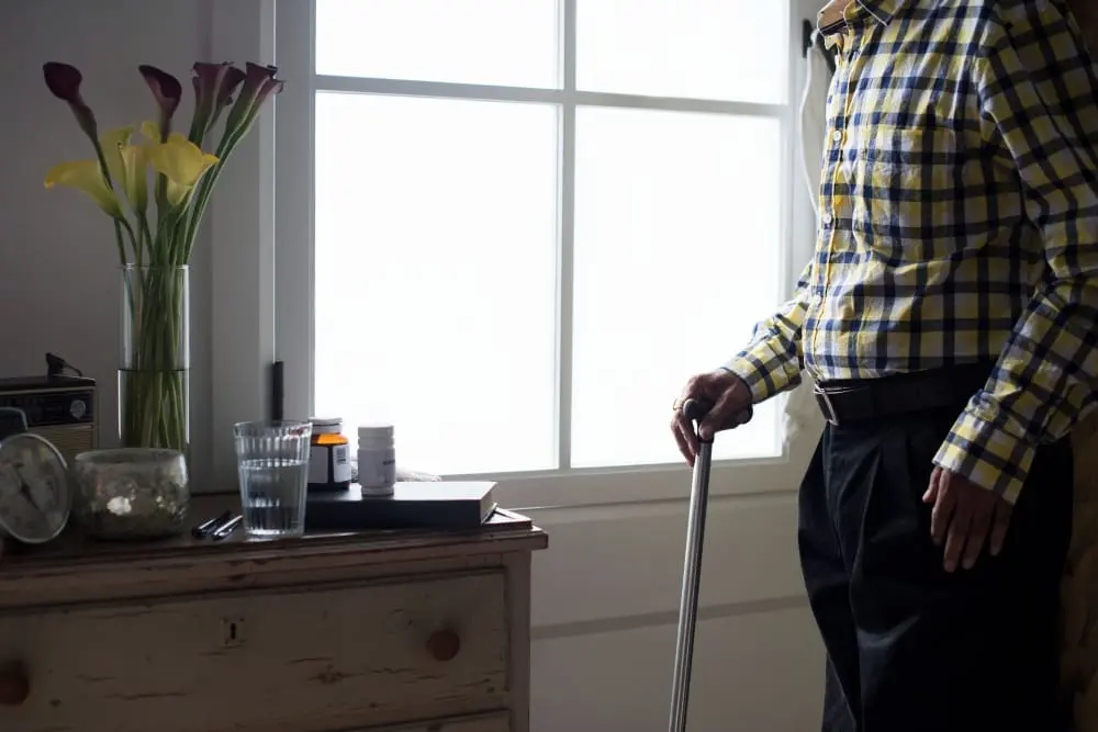 Pessoa idosa usando bengala dentro de casa, com objetos de uso diário próximos.